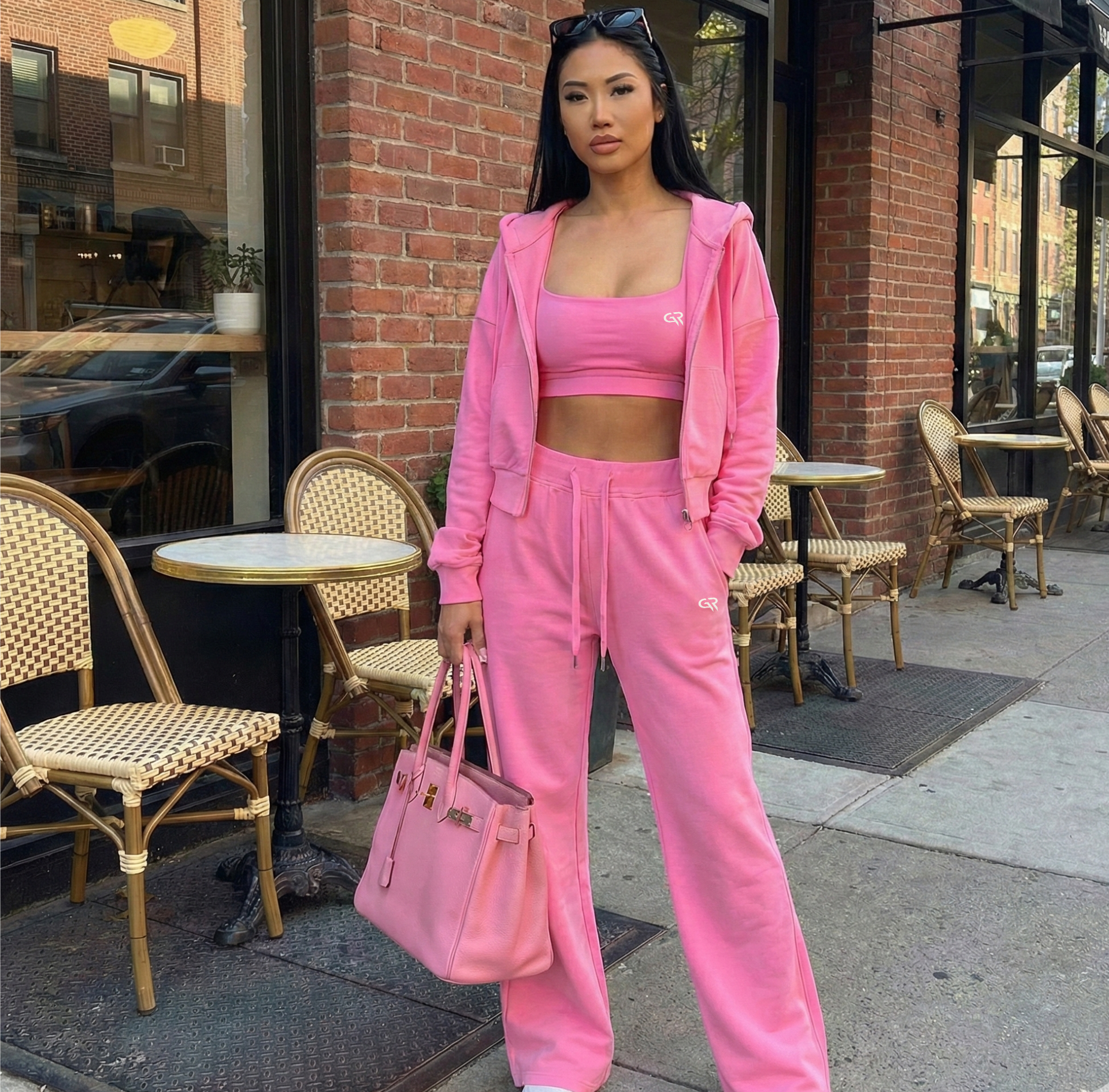 Woman in a pink tracksuit standing on a sidewalk with outdoor cafe tables in the background.